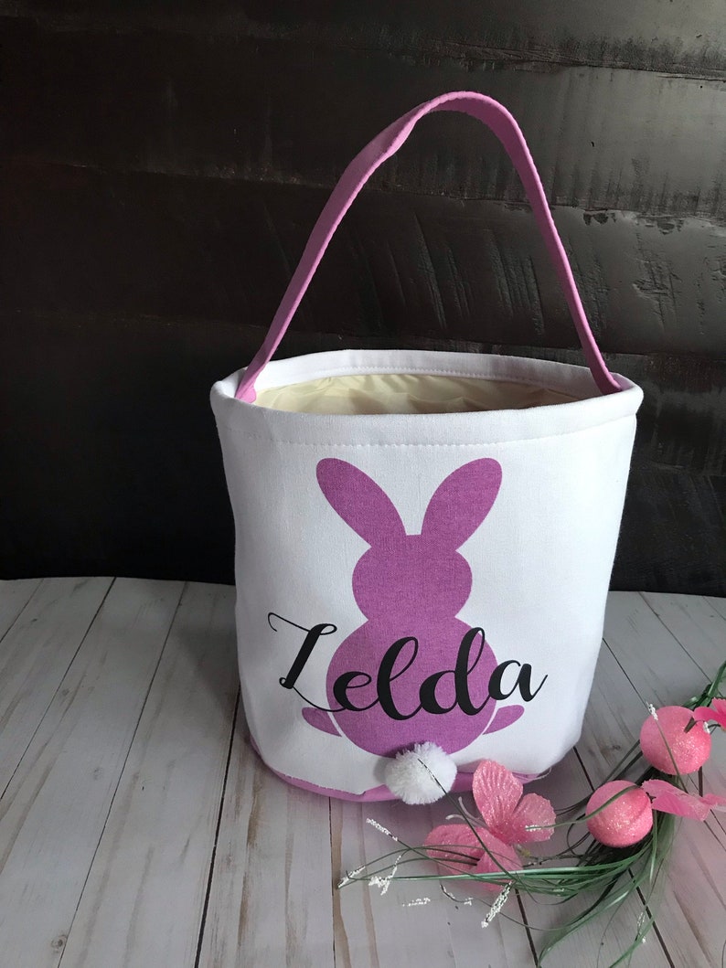 Puede incluir: Una cesta de Pascua blanca con una silueta de conejo morado y el nombre "Zelda" impreso en ella. La cesta tiene un asa rosa y una cola de pomp&oacute;n blanco en el conejo.