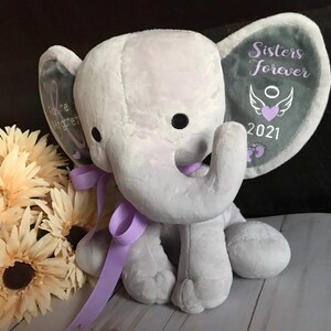 May include: A grey plush elephant with a purple ribbon. The elephant's ears are embroidered with the text "Sisters Forever" and a small angel wing with a heart. The year 2021 is also embroidered on the ear.