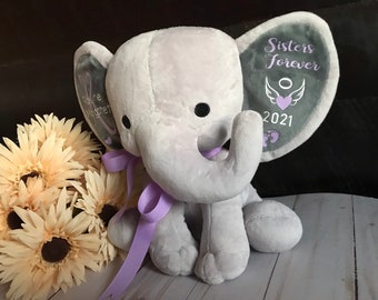 May include: A gray plush elephant with purple ribbon. The elephant's ears are embroidered with the text "Sisters Forever" and a small angel wing with a heart. The year 2021 is also embroidered on the ear.