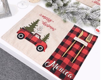 May include: A set of two Christmas-themed placemats. One placemat is beige with a red truck carrying a Christmas tree, and the text "merry christmas". The other placemat is red and black buffalo plaid with the name "Monica" embroidered on it. A gold fork and knife are placed on the plaid placemat.