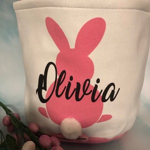 Puede incluir: Cesta de Pascua de lona blanca con una silueta de conejo rosa y el nombre Olivia en letras negras. El conejo tiene una cola de pomp&oacute;n blanco.