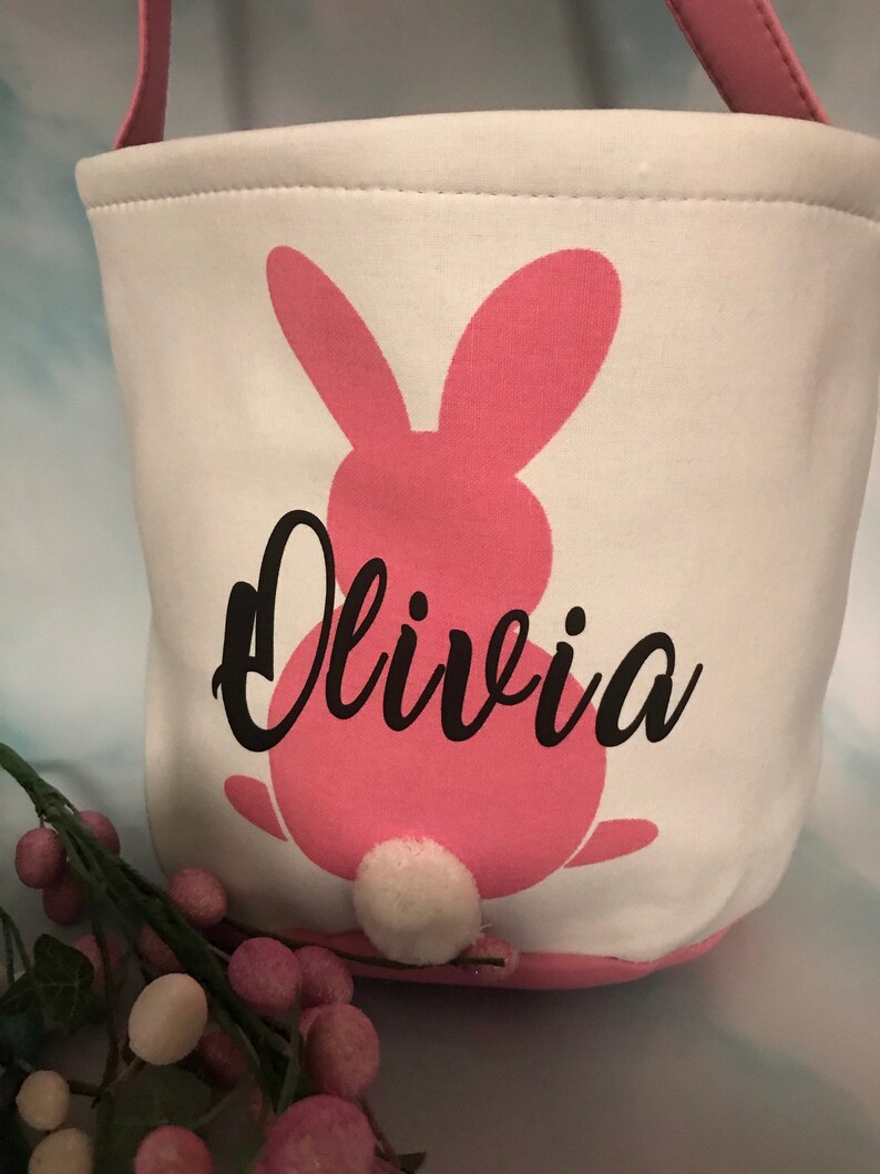 Puede incluir: Cesta de Pascua de lona blanca con una silueta de conejo rosa y el nombre Olivia en letras negras. El conejo tiene una cola de pomp&oacute;n blanco.