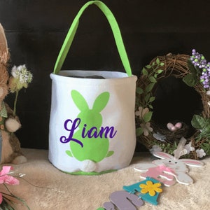Puede incluir: Cesta de Pascua blanca con una silueta de conejo verde y el nombre "Liam" en letras moradas. La cesta tiene un asa verde y est&aacute; sobre una superficie blanca.