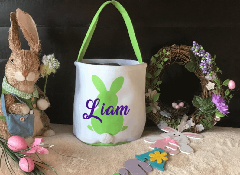 Puede incluir: Cesta de Pascua blanca con una silueta de conejo verde y el nombre "Liam" en letras moradas. La cesta tiene un asa verde y est&aacute; sobre una superficie blanca.
