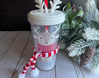 May include: A white tumbler with a reindeer design and a red and white striped scarf. The tumbler has a white lid with a red and white striped straw and white reindeer antlers. The tumbler says "Hello Winter" on the front.
