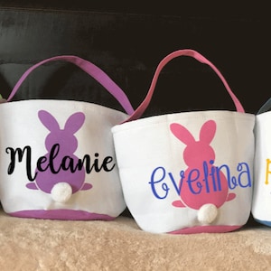 Puede incluir: Cuatro cestas de Pascua de lona blanca con siluetas de conejos y nombres personalizados. Los conejos son verde, morado, rosa y azul. Los nombres son Olivia, Melanie, Evelina y Raizel.