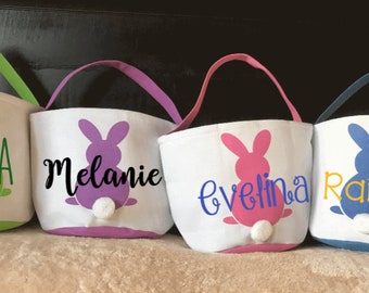 May include: Four white canvas Easter baskets with bunny silhouettes and personalized names. The bunnies are green, purple, pink, and blue. The names are Olivia, Melanie, Evelina, and Raizel.