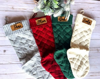 May include: Four knitted Christmas stockings with gold name tags. The stockings are in the colors gray, red, green, and white. The names on the tags are Teresa, Mom, Dad, and Emily.