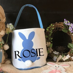Puede incluir: Una cesta de Pascua blanca con una silueta de conejo azul y el nombre "ROSIE" impreso en ella. La cesta tiene un asa azul y una cola esponjosa blanca.