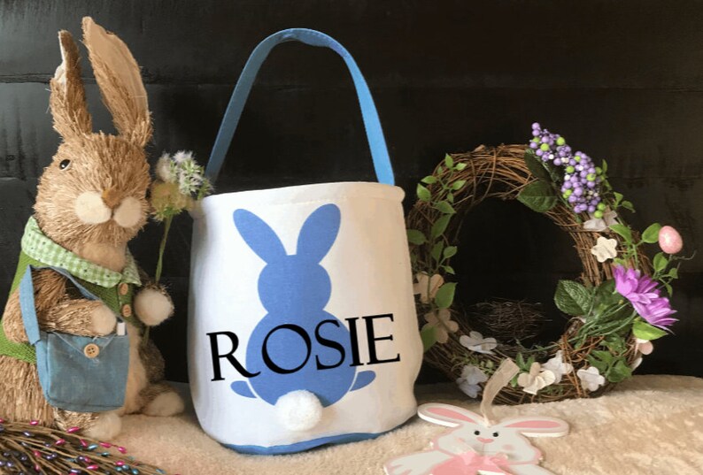 Puede incluir: Una cesta de Pascua blanca con una silueta de conejo azul y el nombre "ROSIE" impreso en ella. La cesta tiene un asa azul y una cola esponjosa blanca.