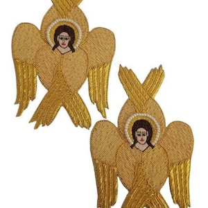 Puede incluir: Dos parches de ángel bordados en oro. Cada parche presenta un rostro detallado con un halo, rodeado de intrincadas alas de hilo dorado y elementos decorativos. Los parches están sobre un fondo blanco.