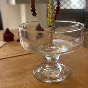 Op de afbeelding: Een heldere glazen schaal op voet met daarin een gebogen draadstructuur versierd met een reeks veelkleurige kralen. De kralen zijn gerangschikt in een volgorde van bruin, lichtgroen, paars, rood, geel en wit voor een kleurrijk geheel.