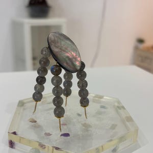 May include: A decorative stand featuring a large, oval, iridescent shell-like centerpiece supported by four legs made of grey, spherical, semi-precious stone beads on gold-toned metal pins, resting on a clear, hexagonal resin tray with embedded stones.