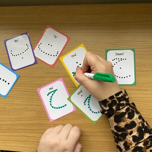Peut inclure: Un ensemble de cartes m&eacute;moire color&eacute;es avec des lettres de l'alphabet arabe &eacute;crites dessus. Chaque carte a une lettre &eacute;crite en points noirs et le nom de la lettre &eacute;crit en texte noir. Une main trace la lettre 'Kha' avec un marqueur vert.