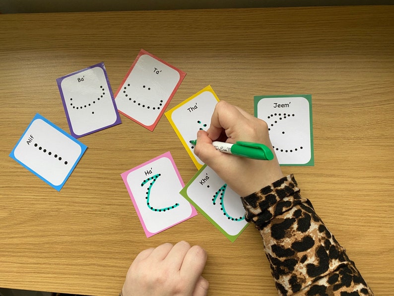 Peut inclure: Un ensemble de cartes m&eacute;moire color&eacute;es avec des lettres de l'alphabet arabe &eacute;crites dessus. Chaque carte a une lettre &eacute;crite en points noirs et le nom de la lettre &eacute;crit en texte noir. Une main trace la lettre 'Kha' avec un marqueur vert.