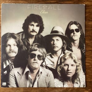 May include: Album cover for Firefall's "Elan." The cover features a sepia-toned photograph of the band members. The band members are wearing various outfits, including sunglasses, hats, and open shirts. The band's name and album title are printed at the top.