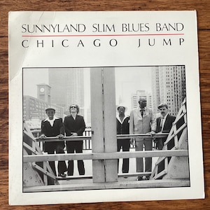 May include: Black and white photo of Sunnyland Slim Blues Band, a group of five men, standing on a bridge in Chicago. The band is performing a live show. The photo is from the album "Chicago Jump".