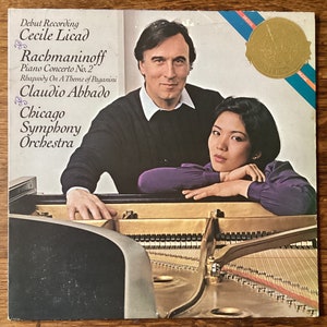 May include: A vinyl record cover featuring a photo of a woman and a man sitting at a grand piano. The cover is for a recording of Rachmaninoff's Piano Concerto No. 2, performed by Cecile Licad and the Chicago Symphony Orchestra, conducted by Claudio Abbado.