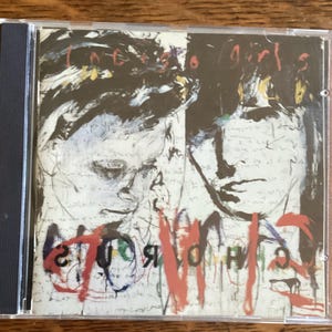 May include: A CD case with a black spine and a clear front cover. The cover art features a black and white abstract painting of two faces with the words "In Every Girls" and "Worlds" written in red and black paint.