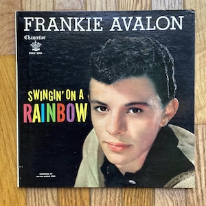 May include: A black and white vinyl record album cover featuring Frankie Avalon. The album title is "Swingin' on a Rainbow" in colourful letters. The album cover features a black and white photo of Frankie Avalon.