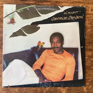 May include: A vinyl record album cover featuring a man in a bright orange shirt sitting on a couch. The album cover is titled "In Flight" by "George Benson". Two small yellow and orange birds are perched on the couch behind the man.