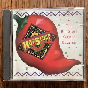 May include: A CD case featuring a large red chili pepper graphic with the words "Hot Stuff" in a stylized logo. The text "The Hot Stuff Catalog Sampler" is also displayed. The CD case is in a clear plastic case.