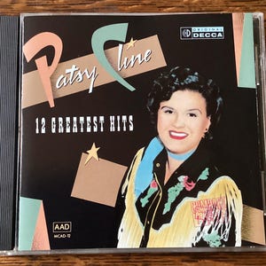 May include: A Patsy Cline CD case with the text "Patsy Cline 12 Greatest Hits." The cover features a portrait of Patsy Cline and a Decca Records logo. The background is black with geometric shapes.