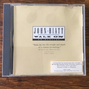 May include: A white CD case with a black and white image of John Hiatt. The text "Walk On CD Sampler" is printed in black on a white background. The text "Limited Edition Promotional Disc includes Bonus Multimedia Track featuring a guide to Hiatt County, lyrics and more. Playable on Mac or Windows. DPRO-10280 Promotional Use Only/Not For Sale" is printed in black on a white background.