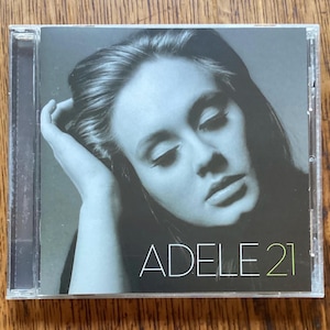 May include: A music CD in a clear plastic jewel case featuring a black and white portrait of a person with their hand near their head. The text ADELE 21 is printed in white and light green at the bottom of the cover art, which is set against a dark background.