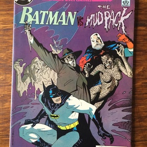 May include: A comic book cover featuring Batman fighting the Mud Pack. The cover is purple and features a dark blue and yellow Batman in a fighting pose. The Mud Pack is a group of villains made up of a green, slimy creature, a woman with brown hair and a green, slimy body, and a man with a blue and white mask.
