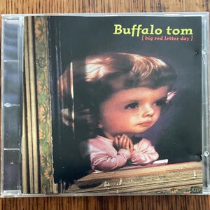 May include: A CD case featuring the album "big red letter day" by Buffalo Tom. The cover art shows a vintage-style illustration of a child with clasped hands, set against a dark background. The band's name is in green text.