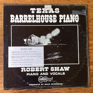 May include: Black and white album cover with a photo of a building with a sign that reads "Barrelhouse Piano". The text "Texas Barrelhouse Piano" is in large white letters at the top of the cover. The text "Robert Shaw Piano and Vocals" is in white letters at the bottom of the cover. The text "Recorded by Mack McCormick" is in white letters at the bottom of the cover. The album cover is for Arhoolie Records, F1010.