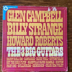 May include: A red vinyl record album cover with the title "The 3 Big Guitars" in yellow letters. The album features Glen Campbell, Billy Strange, and Howard Roberts. The album cover lists the songs "Daddy Roll 'Em", "Sangaree", "Blues Wail", "Cottonfields", "Get the Bird Flyin'", "Nashville Blues Ramble", "Hooten", "Part One", "Bulldurem", "Shindigg'n", and "Color Him Folky".