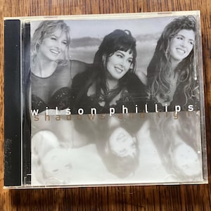 May include: A CD case featuring a black and white image of three women. The text "Wilson Phillips" is above the title "shadows and light". The case is in a clear plastic sleeve with a black spine.