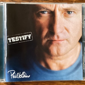 May include: A CD case with a close-up photo of Phil Collins. The album cover is black with white text that reads "TESTIFY" and Phil Collins' signature in white. The background is a blurry image of Phil Collins' face.