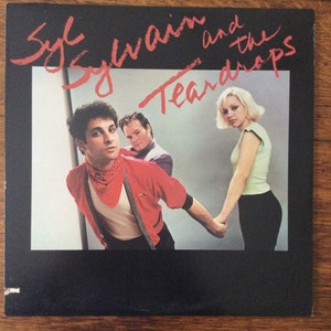 May include: A black and white photo of a band called "Syl Sylvain and the Teardrops". The band members are a man with short brown hair wearing a red sweater and a bandana, a man with short brown hair wearing a blue shirt, and a woman with short blonde hair wearing a green shirt and black pants.