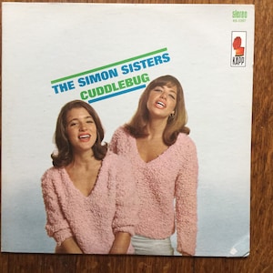 May include: A vintage vinyl record album cover featuring The Simon Sisters. The album cover is white with blue and green text that reads "The Simon Sisters Cuddlebug". The album cover features a photo of two women wearing pink sweaters. The album cover also features a small red and white logo for Kapp Records.