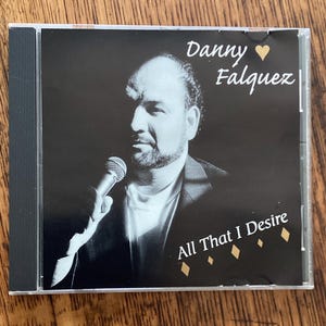 May include: A black and white CD cover with a photo of a man singing into a microphone. The title of the album is "All That I Desire" and the artist is "Danny Falquez".