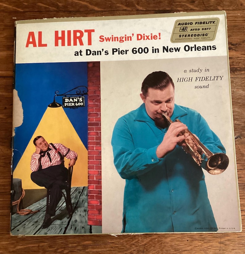 May include: Vintage vinyl record album cover for "Swingin' Dixie!" by Al Hirt. The cover features a man playing a trumpet, and an illustration of a man sitting at a table. The album cover has the text "AUDIO FIDELITY STEREODISC STEREOPHONIC HIGH FIDELITY".