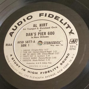 May include: A vintage vinyl record by Al Hirt, featuring his trumpet and Dixieland orchestra, recorded at Dan's Pier 600 in New Orleans. The record label is white with black text, including song titles and timings. The record plays at 33 1/3 RPM.