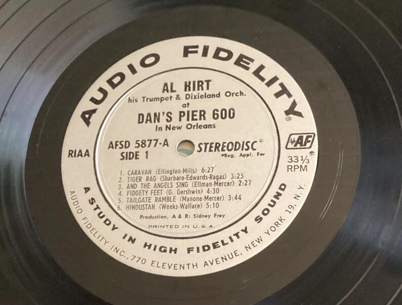 May include: A vintage vinyl record by Al Hirt, featuring his trumpet and Dixieland orchestra, recorded at Dan's Pier 600 in New Orleans. The record label is white with black text, including song titles and timings. The record plays at 33 1/3 RPM.