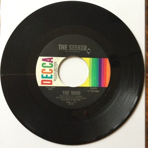 May include: A black vinyl record with a white label. The label has the text "DECCA" in a red, white, and blue font. The text "THE SEEKER (Peter Townshend)" is printed in black. The text "THE WHO" is printed in black. The text "PRODUCED BY KIT LAMBERT" is printed in black. The text "EXECUTIVE PRODUCER CHRIS STAMP" is printed in black. The text "RECORDED IN ENGLAND" is printed in black. The text "(latre: 16) (3:12)" is printed in black. The text "32670 (7-122,560)" is printed in black.
