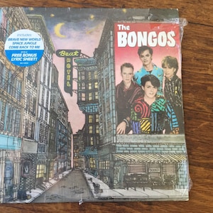 May include: The Bongos album cover features a city street scene with a billboard advertising the band. The billboard is red and white with the band's name in large, white letters. The band members are pictured on the billboard, standing in front of a pink background. The album cover is a vintage style illustration.