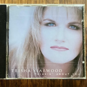 May include: A CD case featuring Trisha Yearwood's album "Thinkin' About You." The cover art showcases a close-up of a woman with blonde hair and blue eyes. The CD case is clear with a black spine.