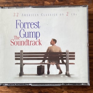 May include: CD case for "Forrest Gump: The Soundtrack" showing a man seated on a bench with a suitcase. The cover displays the title in blue and red, with the text "32 American Classics on 2 CDs".