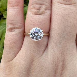 May include: A gold ring with a large, clear, round gemstone. The gemstone is held in place by multiple prongs, and the band is plain gold. The ring is displayed on a finger.