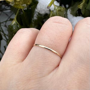 May include: A delicate, thin gold ring. The ring is a simple band design, with a smooth, polished surface. The ring is shown on a finger, with a blurred background of green leaves and snow.