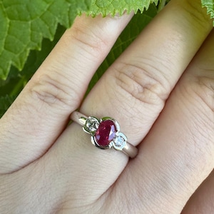 Puede incluir: Un anillo de tono plateado con una piedra preciosa roja central de corte ovalado, flanqueada por dos piedras transparentes más pequeñas de corte redondo. Las piedras están engastadas en una banda de metal liso que rodea el dedo.