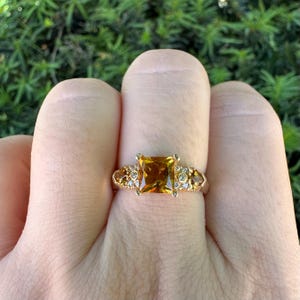 Può includere: Un anello in oro con una grande pietra preziosa color ambra a taglio quadrato al centro, affiancata da pietre chiare rotonde più piccole e ulteriori accenti color ambra incastonati nella fascia, creando un design multi-pietra in oro lucido.