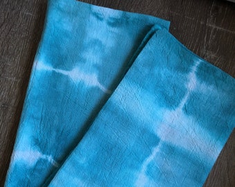 Op de afbeelding: Twee blauwe en witte tie-dye stoffen servetten gevouwen op een donker houten oppervlak.